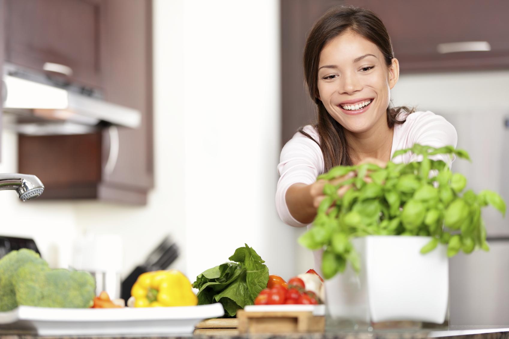 Frau, die Essen in der Küche macht, die nach Basilikum-Anlage greift. Gesundes Essen-Konzept mit der schönen glücklichen lächelnden multi-rassischen kaukasischen / asiatischen Frau zu Hause.